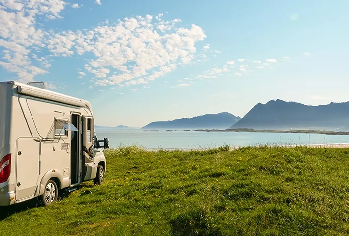 Zijkant van een camper geparkeerd op een grasveld met aan de rechterkant van de foto een meer en bergen in de achtergrond.